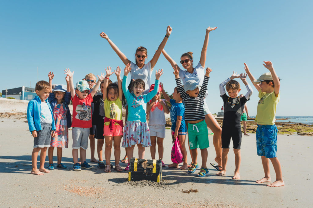 groupes d'enfants sur la plage avec deux moniteurs de voiles, levant les bras au ciel en souriant