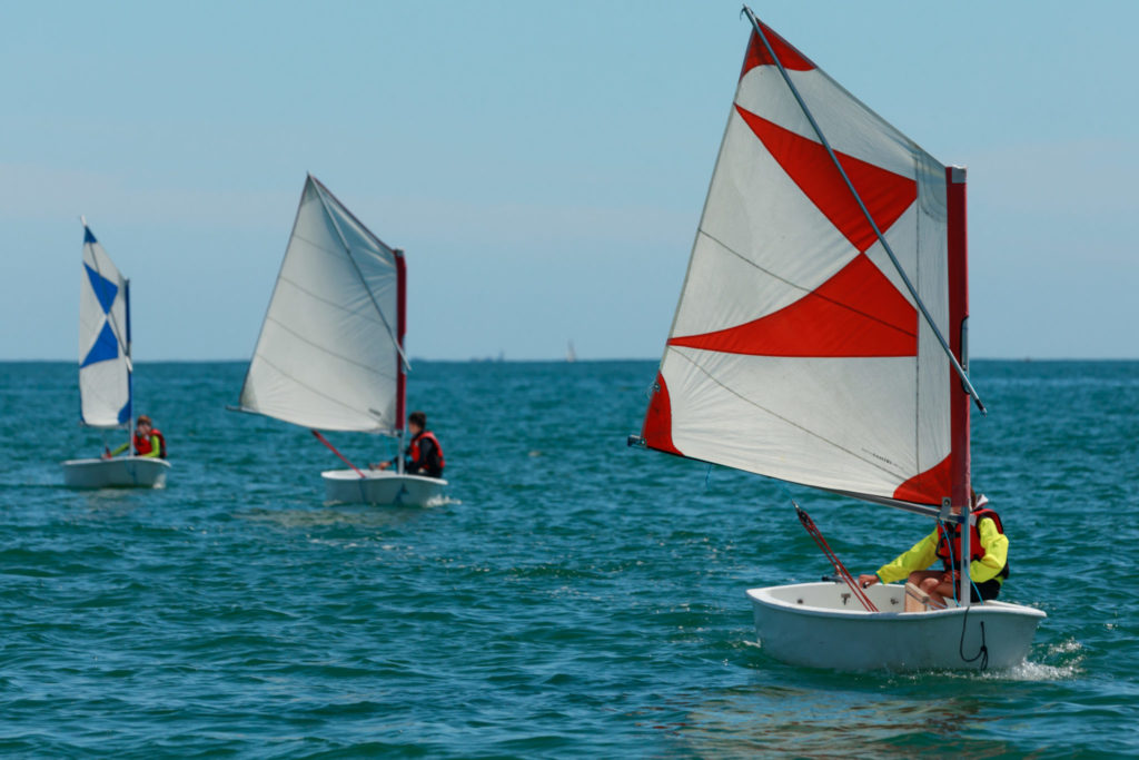 Tois optimist de face, à voiles blanches, bleues et rouges