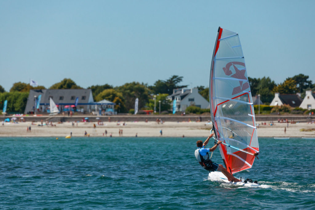 Planche à voile orange naviguant à Loctudy, avec la plage de Langoz en arrière plan